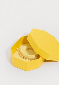 Kollane kuusnurkne kast, mis sisaldab kuldset käevõru, millel on selged kivid ja siidine pael. Kasti sisemus on samuti kollane, millel on tekstuurne viimistlus.