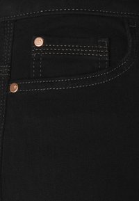 Primo piano di un tessuto in denim nero con dettagli in metallo rame, cuciture bianche e un piccolo taschino. Materiale morbido e testurizzato.