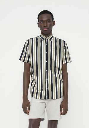 Joven de pie, con una camisa de manga corta abotonada con rayas verticales negras, blancas y oliva, y pantalones cortos beige claro.