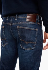 Tmavě modré džínové kalhoty s hnědou koženou záplatou na zadní straně. Vyznačují se viditelným prošitím kapes s detailním vzorem ve tvaru diamantu.