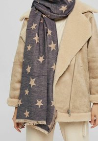 Veste en shearling beige avec grands revers, associée à une écharpe gris foncé à motif d'étoiles et un pantalon crème côtelé.