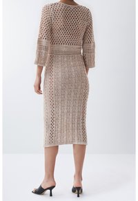 Robe en maille beige clair avec motifs ajourés, manches trois-quarts et coupe ajustée, se terminant sous le genou.