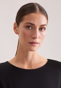 Czarna sukienka z okrągłym dekoltem, gładką teksturą i dopasowanym krojem, noszona przez osobę z zaczesanymi do tyłu włosami i minimalnym makijażem.