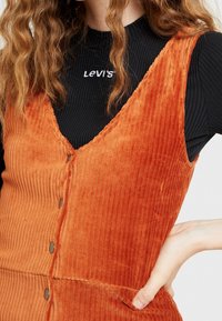 Vestito in corduroy arancione con scollatura a V profonda, caratterizzato da bottoni frontali, texture a coste e una silhouette aderente. Top nero a coste sotto.