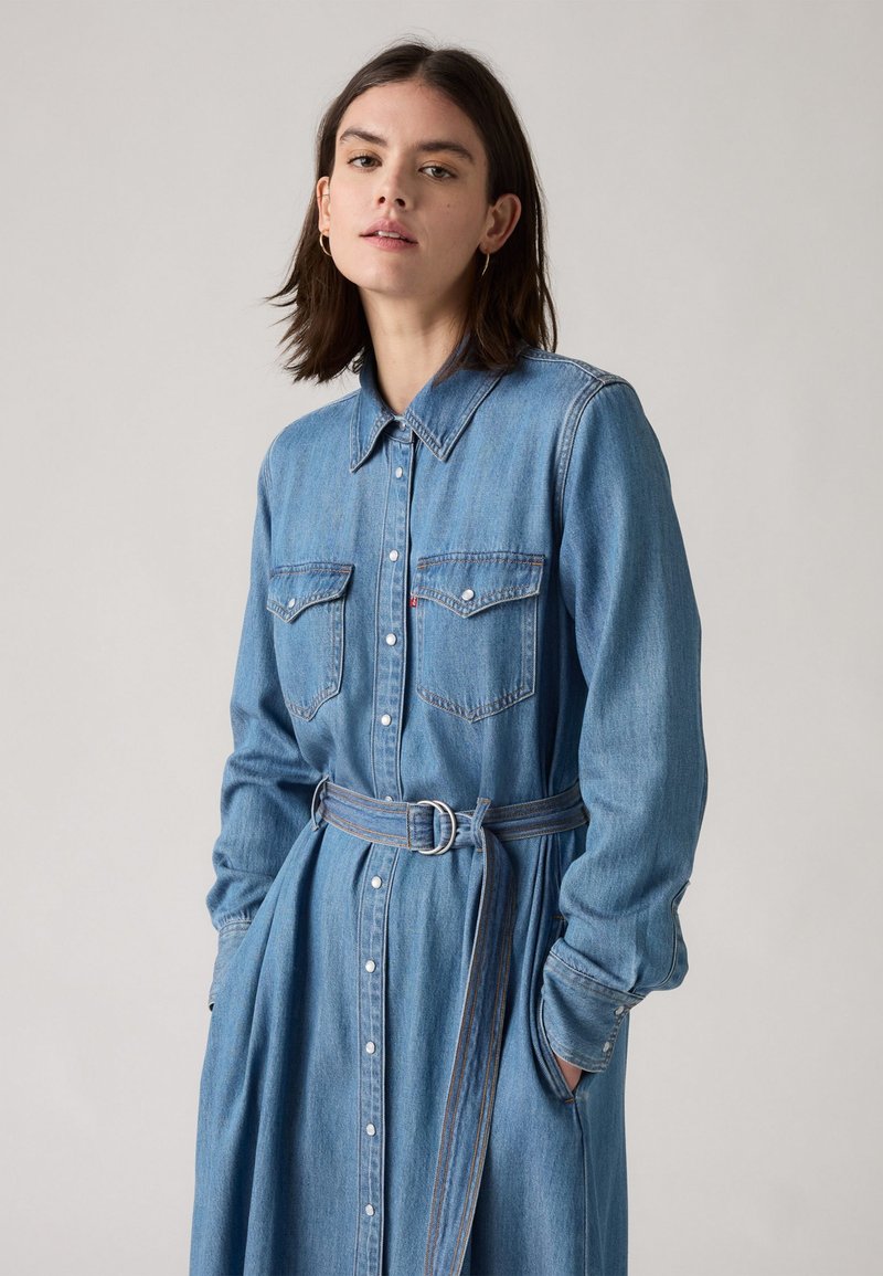 Robe chemise en denim bleu clair. Présente des manches longues, un col pointu, deux poches poitrine et une ceinture nouée à la taille avec une boucle argentée.