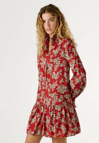 Rode bloemenjurk met lange mouwen, knopen aan de voorkant en geplooide rok. Heeft een beige bloemenpatroon op lichte stof.