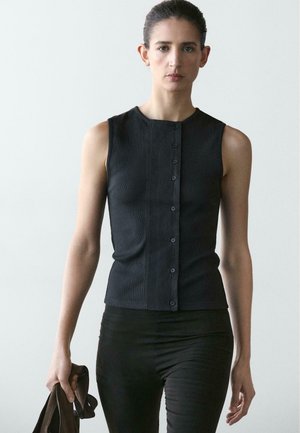 Femme aux cheveux foncés portant un haut noir sans manches boutonné et un pantalon noir, marchant vers l'avant en tenant une veste marron dans sa main gauche.