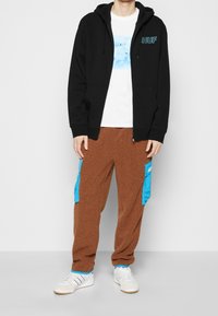 Felpa nera con zip e logo "HUF", t-shirt bianca con grafica, e pantaloni di pile marroni con tasche cargo blu. Completa il look con sneakers bianche.