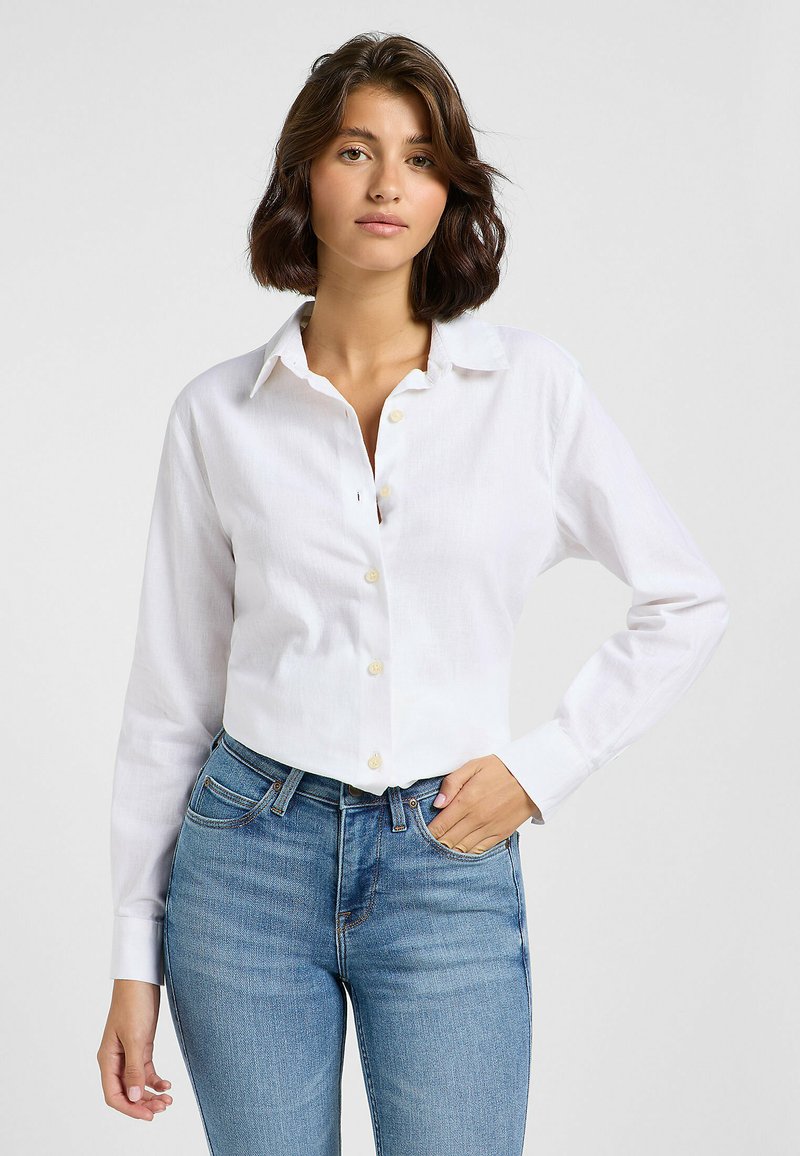 Weißes, kurzes Blusenoberteil mit langen Ärmeln, Knopfverschluss vorne und kragenförmigem Ausschnitt, kombiniert mit hellblauen Jeans. Glatte Textur, minimalistisches Design.