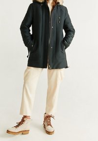 Ciemnozielona kurtka parka z kapturem i sztucznym futerkiem, z zamkiem z przodu, bocznymi kieszeniami i luźnym krojem, noszona na jasnobeżowych spodniach.