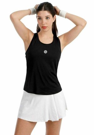 Jonge vrouw in een zwarte mouwloze top en een witte rok die haar haar met opgetrokken armen tegen een witte achtergrond aanpast.