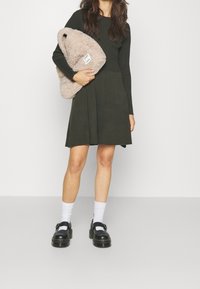 Mörkgrön stickad klänning med ribbad textur, utställd kjol och långa ärmar. Håller en beige fluffig väska med en etikett, bär svarta skor och vita strumpor.