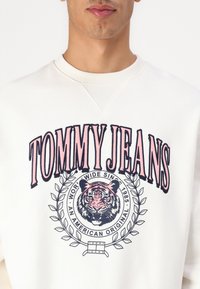 Creme sweatshirt met een grote gekleurde afbeelding van een tijger, tekst "TOMMY JEANS" in roze en zwart, met een cirkelvormig laurelontwerp.