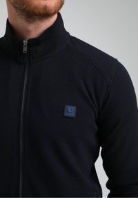 Giacca zip-up blu navy realizzata in tessuto strutturato, con colletto alto e una patch con logo blu sul lato sinistro del petto.