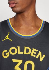 Sort basketballtrøje med gul tekst "GOLDEN" og nummeret "30" i hvid. Blå og gul kant langs halsudskæringen med et Jordan-logo.