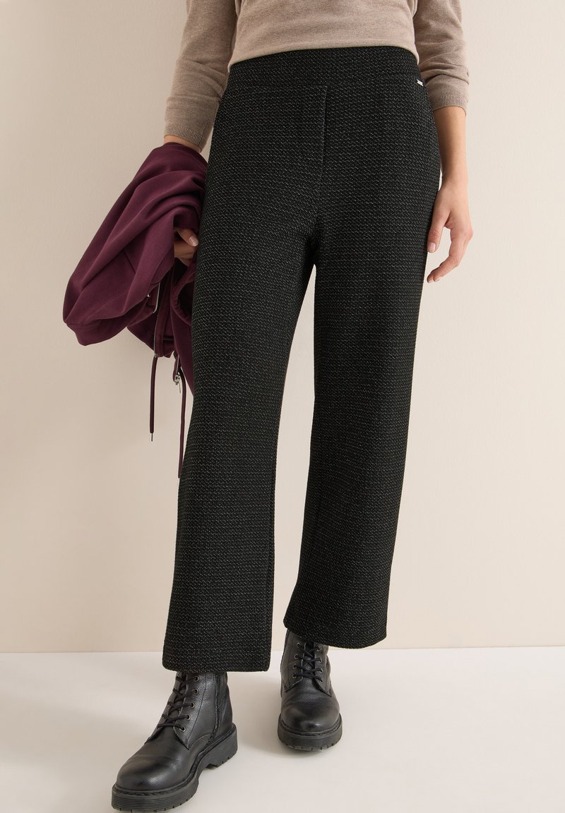 Pantalons larges noirs à texture avec taille haute, associés à une veste bordeaux tenue à la main et des bottes à lacets noires.