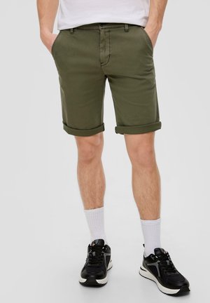 Olivgrüne Shorts aus leichtem Stoff, mit umgeschlagenen Säumen, Seitentaschen und einem Reißverschluss vorne. Getragen mit schwarzen Sneakers und weißen Socken.
