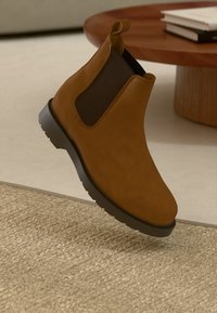 Bota Chelsea castanha com superfície em couro liso, painéis laterais elásticos, uma alça de puxar e sola de borracha texturizada, elevada acima de um tapete em fibras.