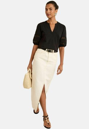 Frau trägt eine schwarze Bluse mit Spitzensäumen, einen cremefarbenen asymmetrischen Rock mit einem vorderen Schlitz, schwarze Sandalen und hält eine gewebte Handtasche.