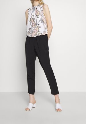 Femme portant un haut blanc sans manches avec un motif paisley rose, un pantalon noir fuselé et des mules blanches à talons ouverts, debout devant un fond uni.