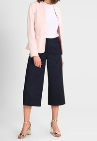 Světlé růžové střižené sako, bílé tričko, námořnické široké kalhoty a zlaté sandály na blokovém podpatku. Hladké textury a strukturovaný design.