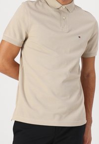 Beige poloko s texturovanou tkaninou, límečkem, třemi knoflíky a malým logem na levé části hrudi. Krátké rukávy s žebrovanými manžetami.