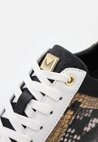 Gros plan d'une basket noire et blanche avec des lacets blancs, un œillet doré, une languette noire texturée et un logo Michael Kors sur le panneau latéral à motif peau de serpent.
