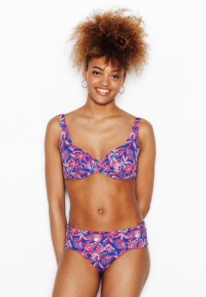 Femme souriante en bikini bleu et rose à motif floral, avec de grands cheveux bouclés et des boucles d’oreilles anneaux, debout devant un fond blanc.