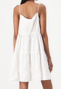 Biała bawełniana sukienka letnia z regulowanymi cienkimi ramiączkami, warstwowym fasonem i marszczonym pasem. Posiada okrągły dekolt z tyłu.