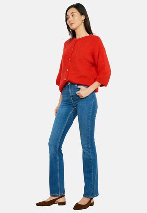 Donna che indossa un cardigan rosso acceso con bottoni, jeans blu a gamba dritta e scarpe marroni con tacco basso, in piedi con una mano in tasca.