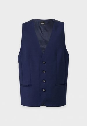 Un homme porte un gilet bleu marine boutonné sur une chemise bleu clair, avec un pantalon foncé, une ceinture beige et des chaussures de ville marron.