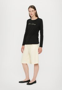 Black long-sleeve shirt with gold script text and cream-colored shorts. Model wears black flats. Simple, fitted design. Smooth fabric texture.