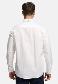 Homme portant une chemise blanche à manches longues boutonnée et un pantalon sombre, debout dos à la caméra sur un fond uni.