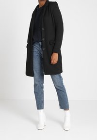 Cappotto lungo nero con un design strutturato, sei bottoni e tasche laterali, abbinato a jeans azzurri chiari e stivali bianchi alla caviglia.