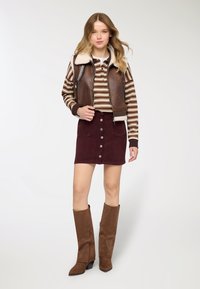 Gilet in pelle marrone con colletto in shearling, camicia a righe con maniche lunghe, gonna in velluto a coste bordeaux con bottoni e stivali marroni al ginocchio.