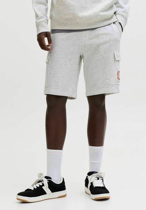 Personne portant un short cargo gris clair, des chaussettes blanches montantes et des baskets noires à lacets blancs, debout devant un fond uni.