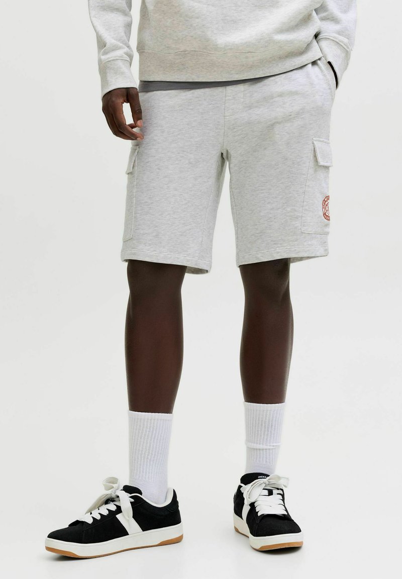 Osoba v svetlosivých cargo krátkych nohaviciach, bielych členkových ponožkách a čiernych teniskách s bielymi šnúrkami, stojaca pred jednoduchým pozadím.