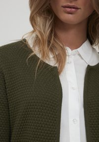 Olivově zelený texturovaný cardigan s kulatým límcem, který je spárován s bílou košilí na knoflíky. Pohled zblízka se zaměřuje na detaily látky a designu.