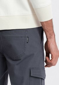 Grå cargo bukser med sidelomme og baglomme, lavet af tykkere stof. Hvide sweatshirt ærmer synlige for oven. Et mærke hænger fast på bukserne.