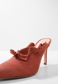 Rostfarbene Wildleder-High-Heel-Mule mit spitzer Zehenpartie und Rüsche am oberen Rand auf einem weißen Hintergrund.