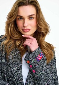 Mladá žena s vlnitými svetlohnedými vlasmi, oblečená v vzorovanom saku s farebnými gombíkmi a bielou blúzkou, ktorá si opiera ruku neďaleko brady.