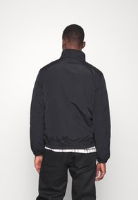 Man som bär en svart pufferjacka, svarta byxor och en rutig skjorta, står vänd mot en vit vägg med avslappnade händer vid sidorna.