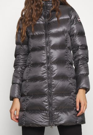 Femme portant une longue doudoune matelassée brillante de couleur gris foncé avec fermeture éclair à l'avant et manches longues, debout devant un fond uni.