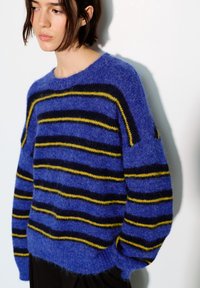 Pull en maille bleu avec des rayures horizontales noires et jaunes. Texture douce avec une coupe décontractée, poignets et ourlet côtelés.