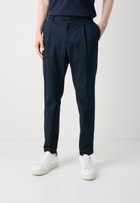 Marinblå veckade byxor med skräddarsydd passform, tillverkade i ett mjukt tyg. Kombinerade med vita sneakers för en avslappnad look.