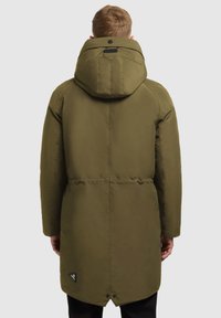 Olīvu zaļš parka ar kapuci, strukturēta ārējā auduma, un logo akcentu aizmugurē. Iespējo sasprindzinātu vidukli un pogu aizdari apakšmalā.