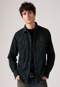 Camisa de mezclilla verde oscuro con botones, dos bolsillos en el pecho, mangas largas y botones a presión, superpuesta sobre una camiseta negra.