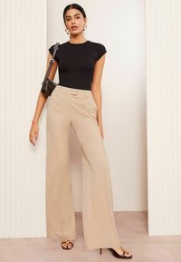 Schwarzes, figurbetontes Oberteil und beigefarbene, weit geschnittene Hosen. Die Hose hat eine strukturierte Taille und Vorderfalten. Schwarze Handtasche und Riemchensandalen.