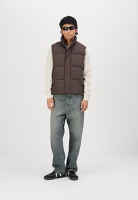 Brun quilted puffer vest over en lys grå sweatshirt, parret med løse blå jeans og sorte sneakers med hvide detaljer. Solbriller båret.