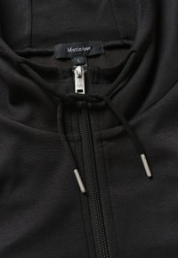 Sweat à capuche noir zippé avec cordons, tirette argentée marquée d'un "M" et étiquette de vêtement mentionnant "Matinique L."